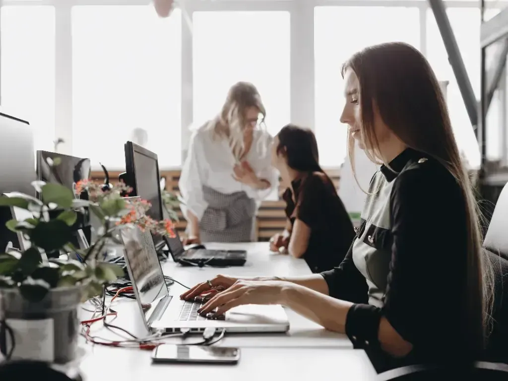 Duas profissionais trabalham em computadores junto a uma mesa decorada com plantas.