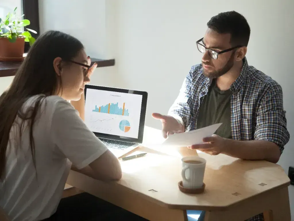Mulher e homem analisam juntos gráficos exibidos em um monitor sobre a mesa.