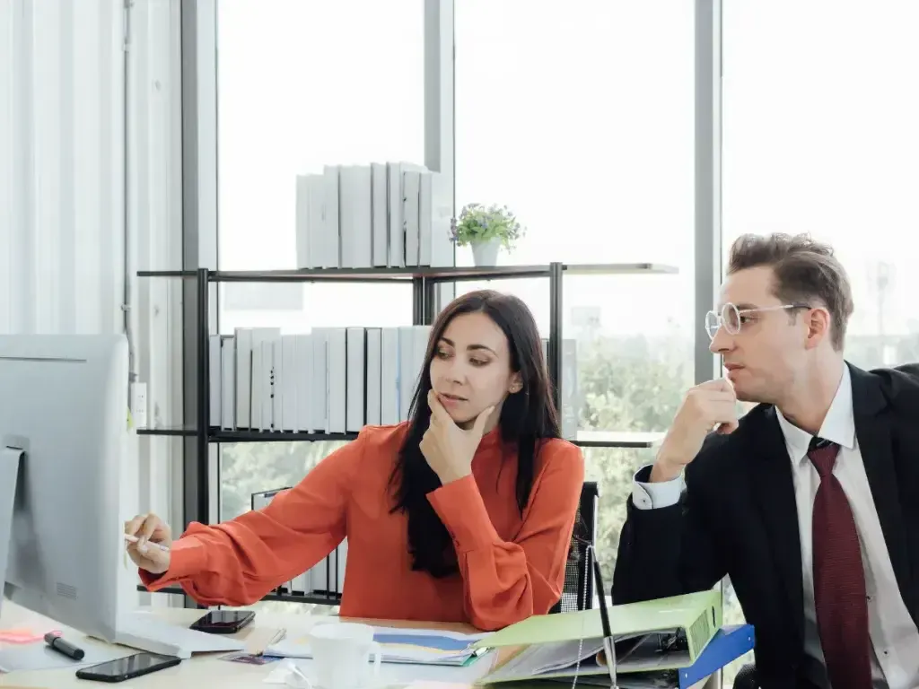 Mulher gesticula enquanto conversa com colega diante de documentos e notebook.