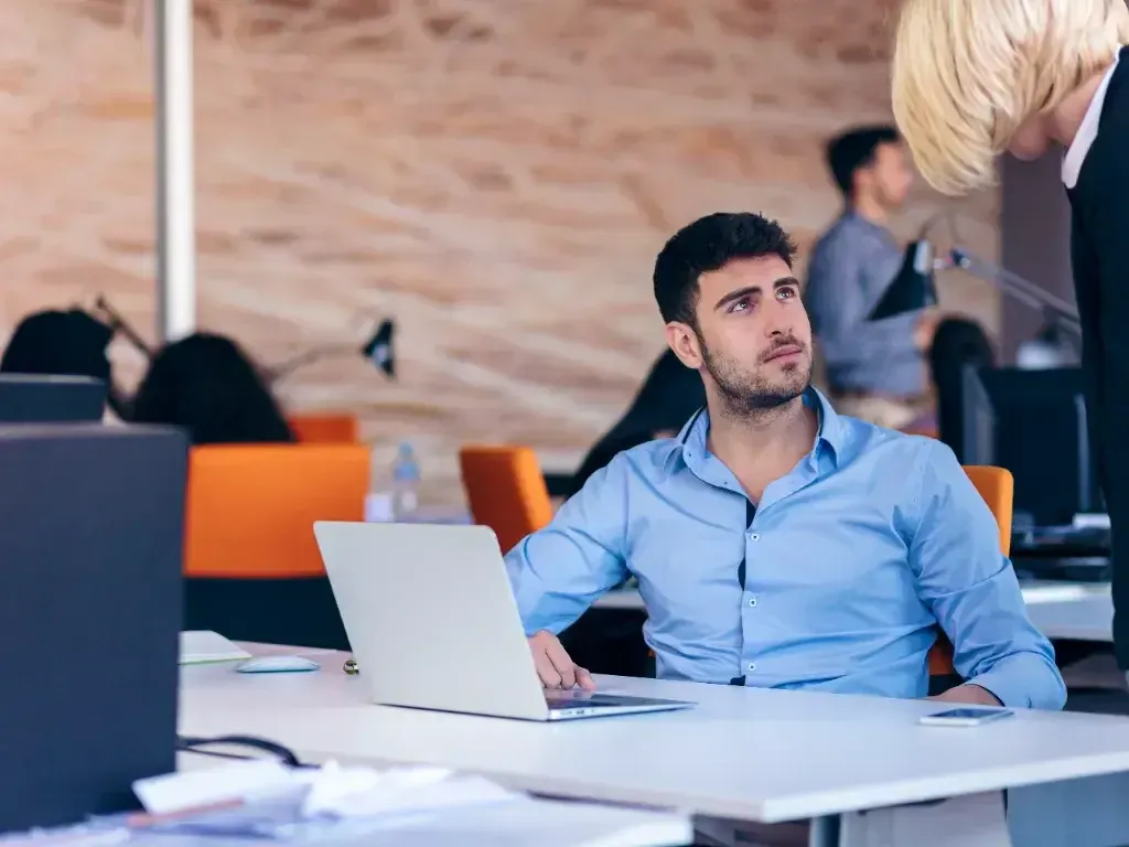 Homem usa notebook em uma mesa de escritório, com colegas ao fundo.