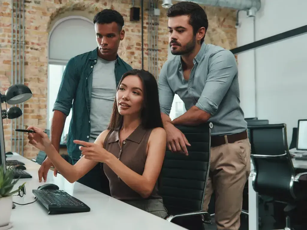 Três colegas observam um monitor enquanto uma mulher aponta para a tela.