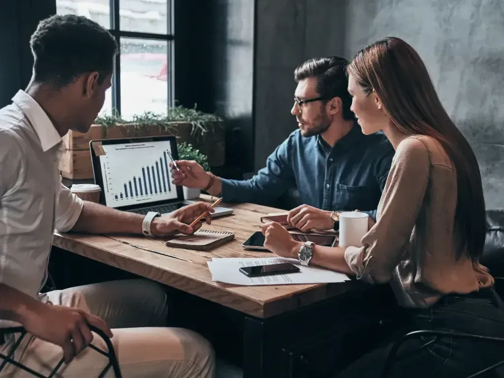 Três profissionais analisam um notebook e documentos durante uma reunião de trabalho.