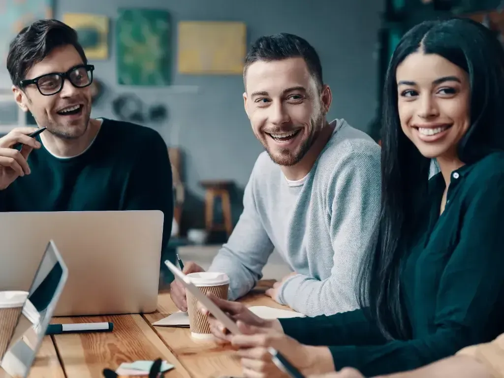 Três colegas sorridentes conversam ao redor de um notebook em uma mesa.