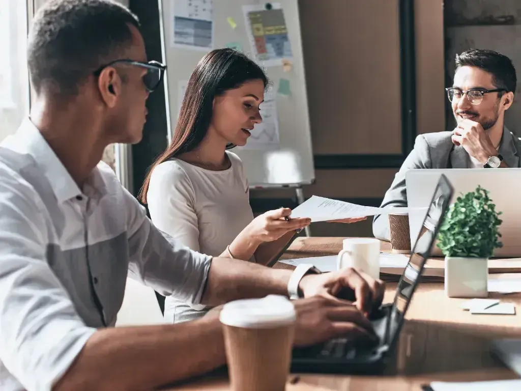 Três profissionais conversam e analisam documentos ao redor de um notebook na mesa.
