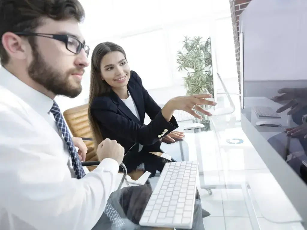 Dois profissionais sentados à mesa analisam juntos a tela de um computador.
