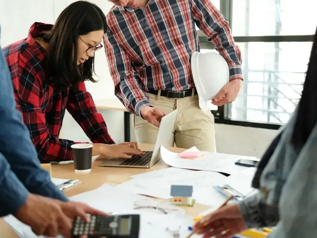 Pessoas analisam documentos e uma calculadora ao redor de uma mesa de trabalho.