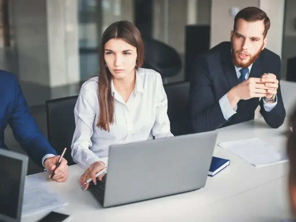Mulher e homem conversam diante de um notebook durante uma reunião.