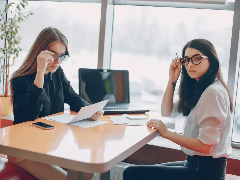 Duas mulheres conversam e analisam papéis diante de um notebook.