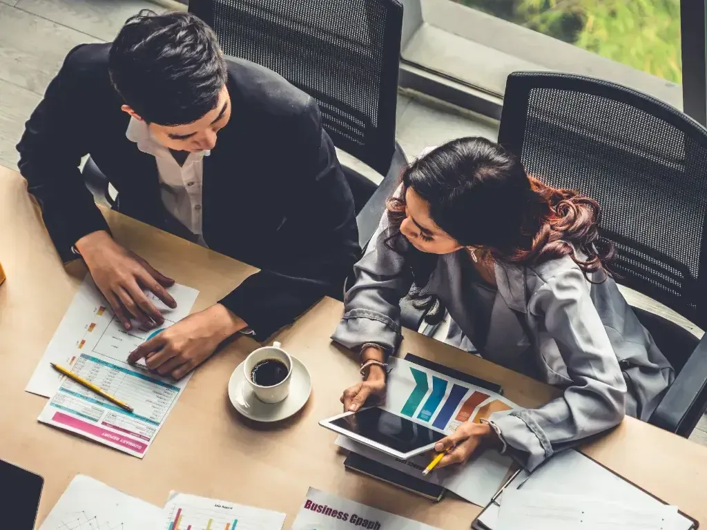 Duas pessoas observam relatórios e gráficos espalhados sobre uma mesa.