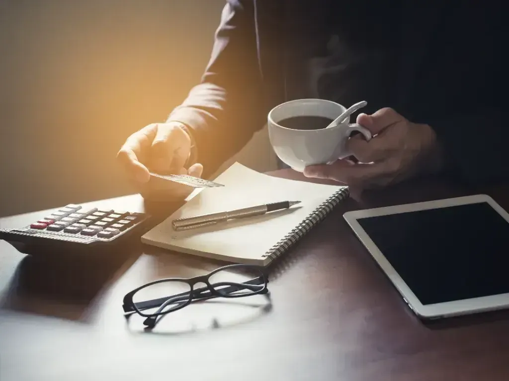 Mão usa uma calculadora ao lado de óculos, papéis e tablet sobre a mesa.